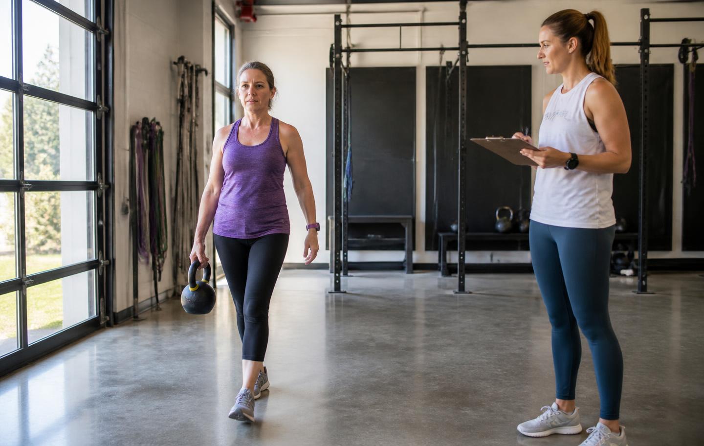 A woman in her mid fifties carrying a kettlebell in a training studio while her coach records her progress